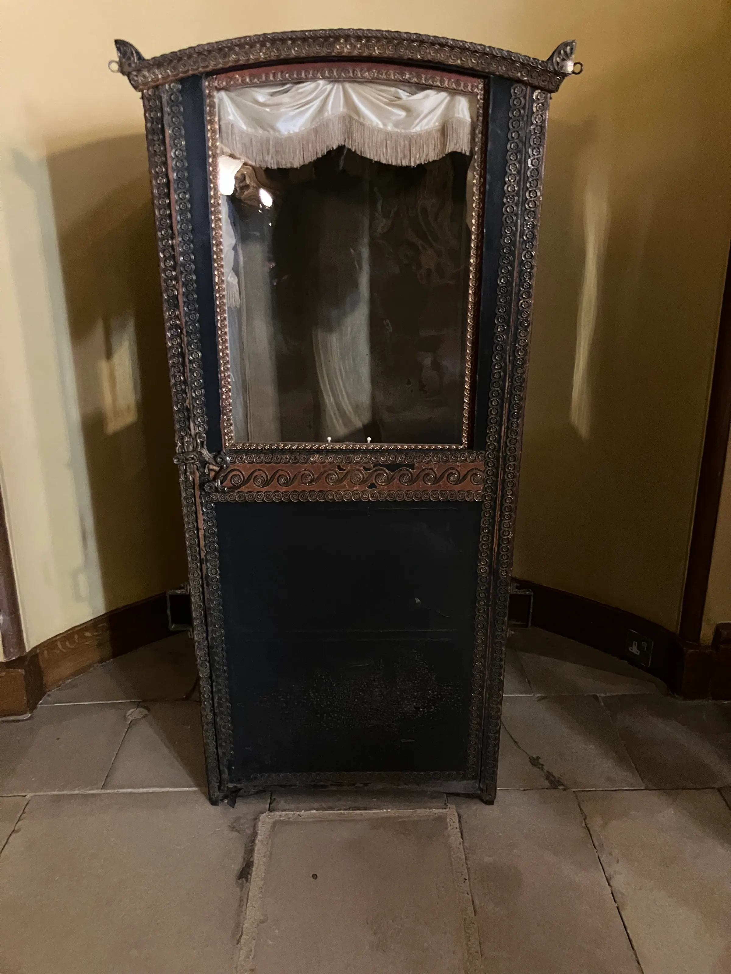 Looks like an old time telephone booth. No this is a chair that a person sat in and then the servants, using poles on each side, carried the person from one place to another.